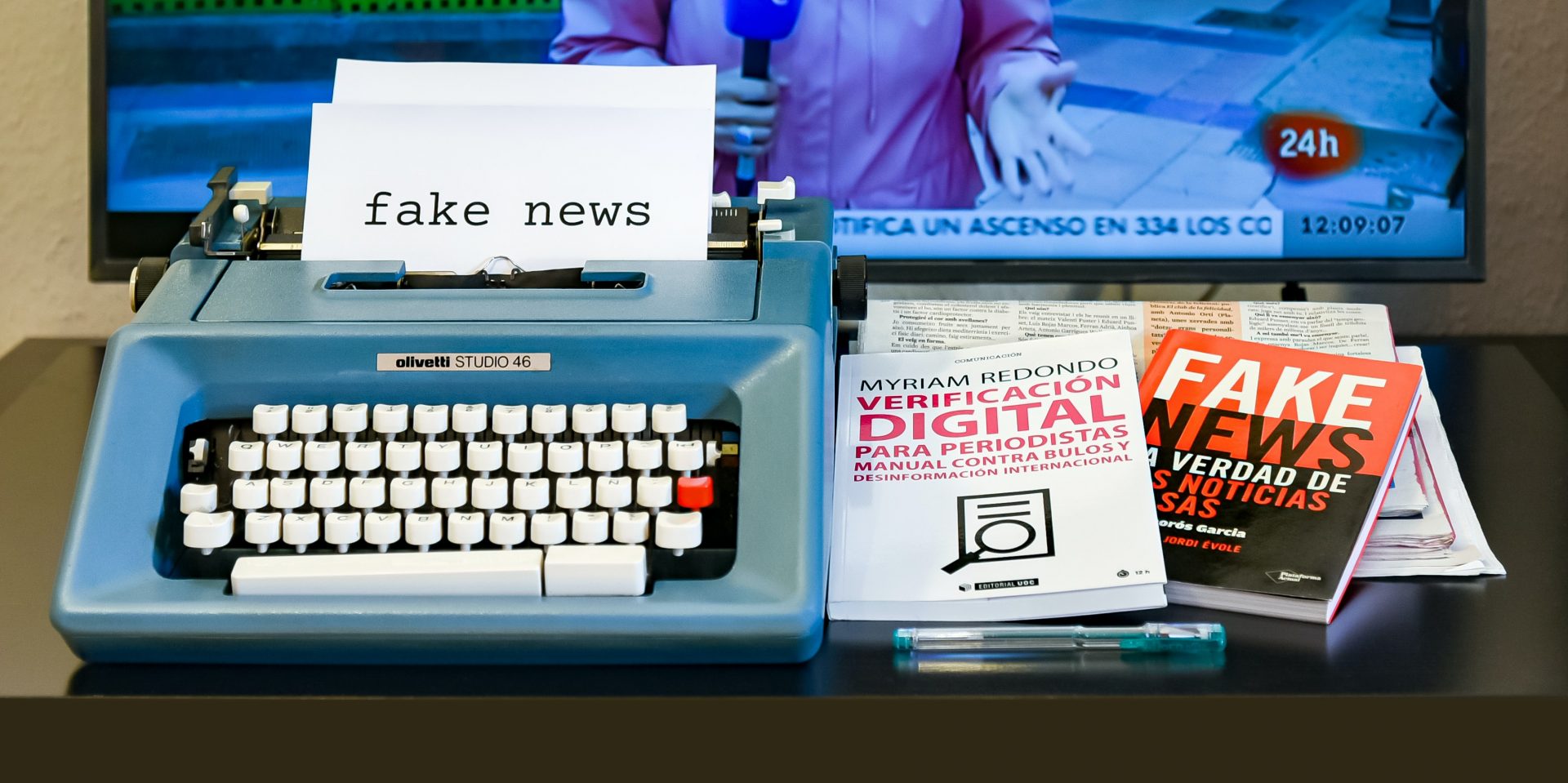 Typewriter with printed paper reading fake news along with a pen and two books on top of newspapers and a television screen behind