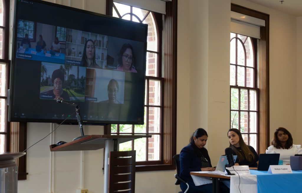 Robyn Caplan, Nanditha Narayanamoorthy, and Syeda Zainab Akbar Lnu talking during Panel 2, with the virtual panelists featured on TV.