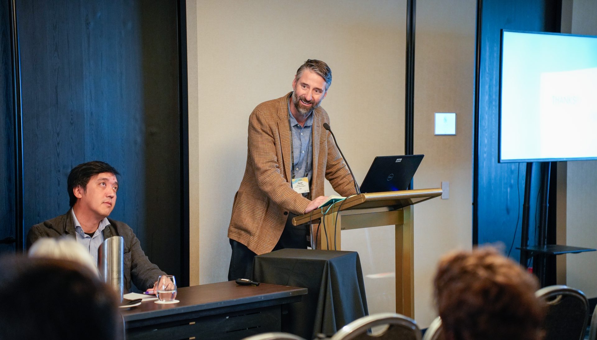 A man with a beard and a brown coat speaking at a lectern, addressing an audience in a room. There is a notebook and a laptop on the lectern. The man is speaking into a microphone. Another man with a gray coat and a light blue button-down is seated at a table next to the lectern with a glass of water, listening attentively.