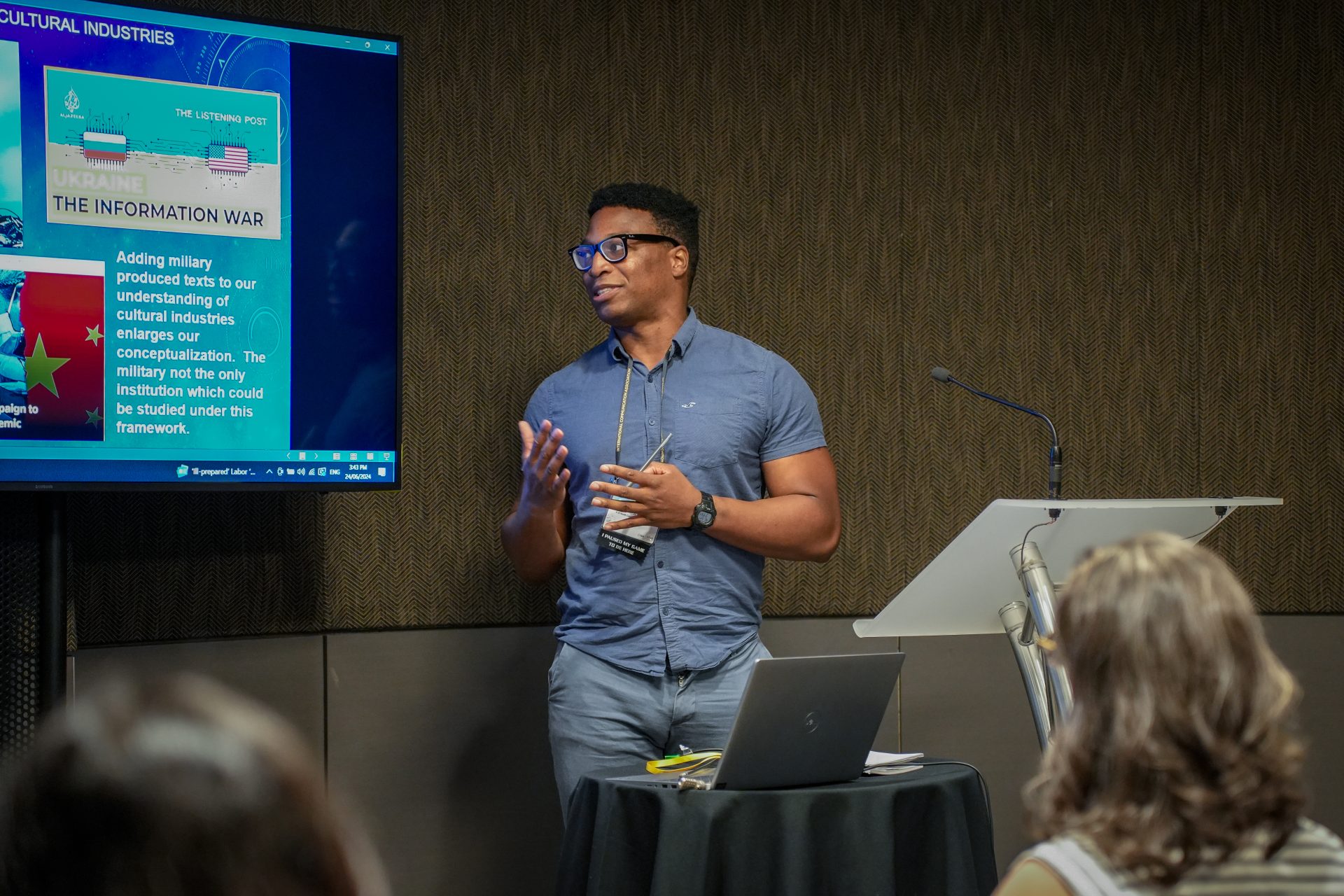 A man with glasses stands at a lectern while giving a presentation addressing an audience of people. He gestures to the screen while speaking.