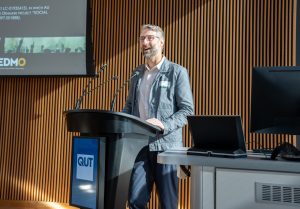 A bearded man wearing glasses engages an audience in a room, with a presentation displayed behind him.