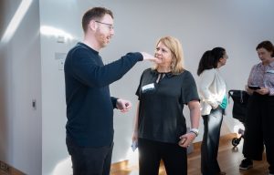 A man with glasses and a navy long-sleeve top conversing with a woman with straight hair and a black top, with two other women engaged in conversation in the background.