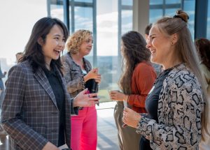 Two women, one with her hair down and one with half-up, half-down hair, conversing, with two other women in the background.