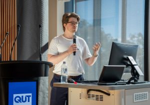 A man with glasses holding a microphone, giving a presentation by a lectern to an audience.