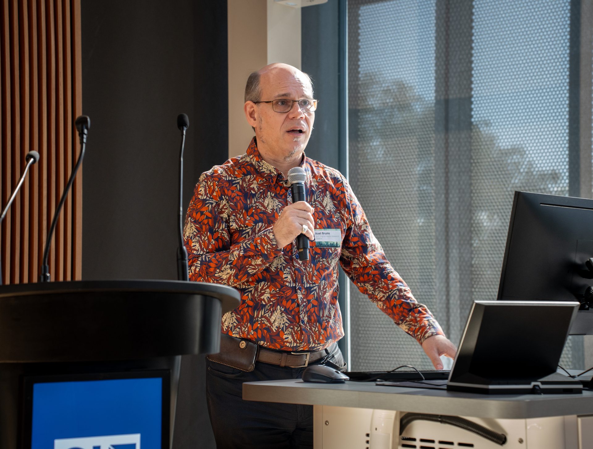 A man with glasses, wearing a colorful shirt, holding a microphone.