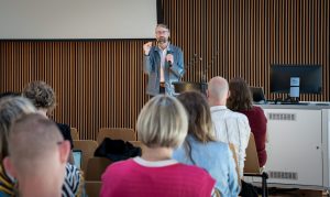 A man with glasses, wearing a coat, holding a microphone and giving a presentation by a lectern to an audience.