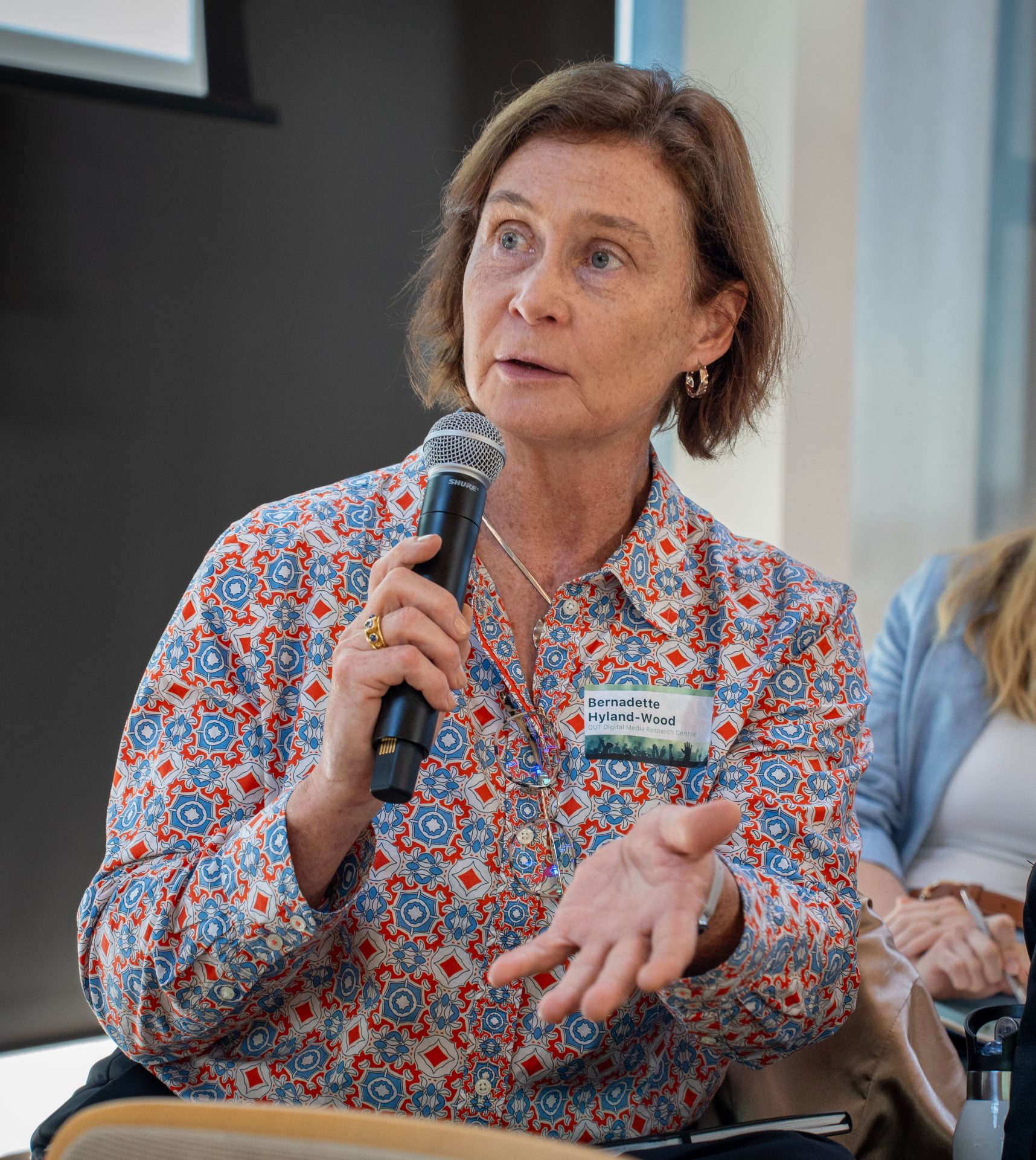 A woman wearing a colorfully patterned button-down shirt, holding a microphone.