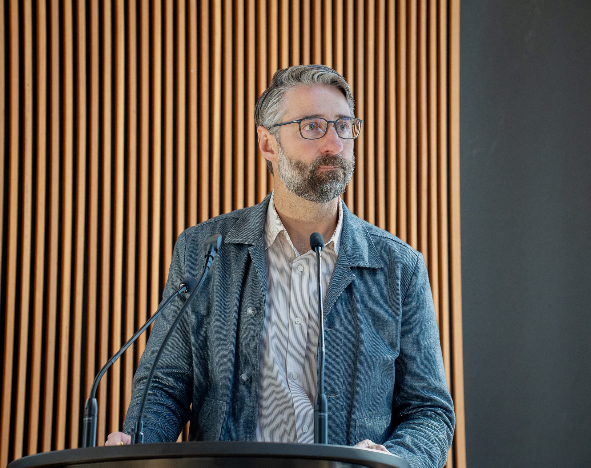 A man with a beard and glasses standing at a lectern, looking attentive.