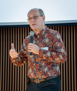 A man wearing a colorful button-down shirt, holding a microphone and speaking.