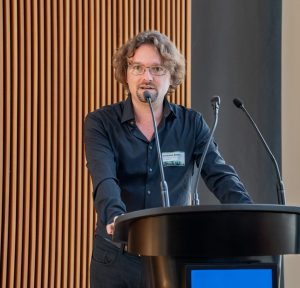 A man with glasses and a black button-down shirt speaking into a microphone by a lectern.