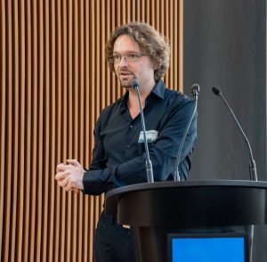 A man with glasses and a black button-down shirt speaking into a microphone by a lectern.