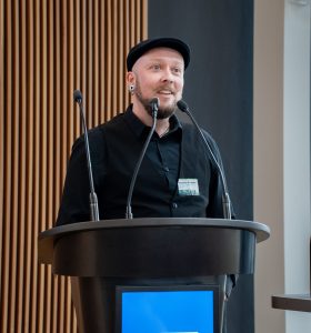 A man with a black cap and a black button-down shirt speaking into a microphone by a lectern.