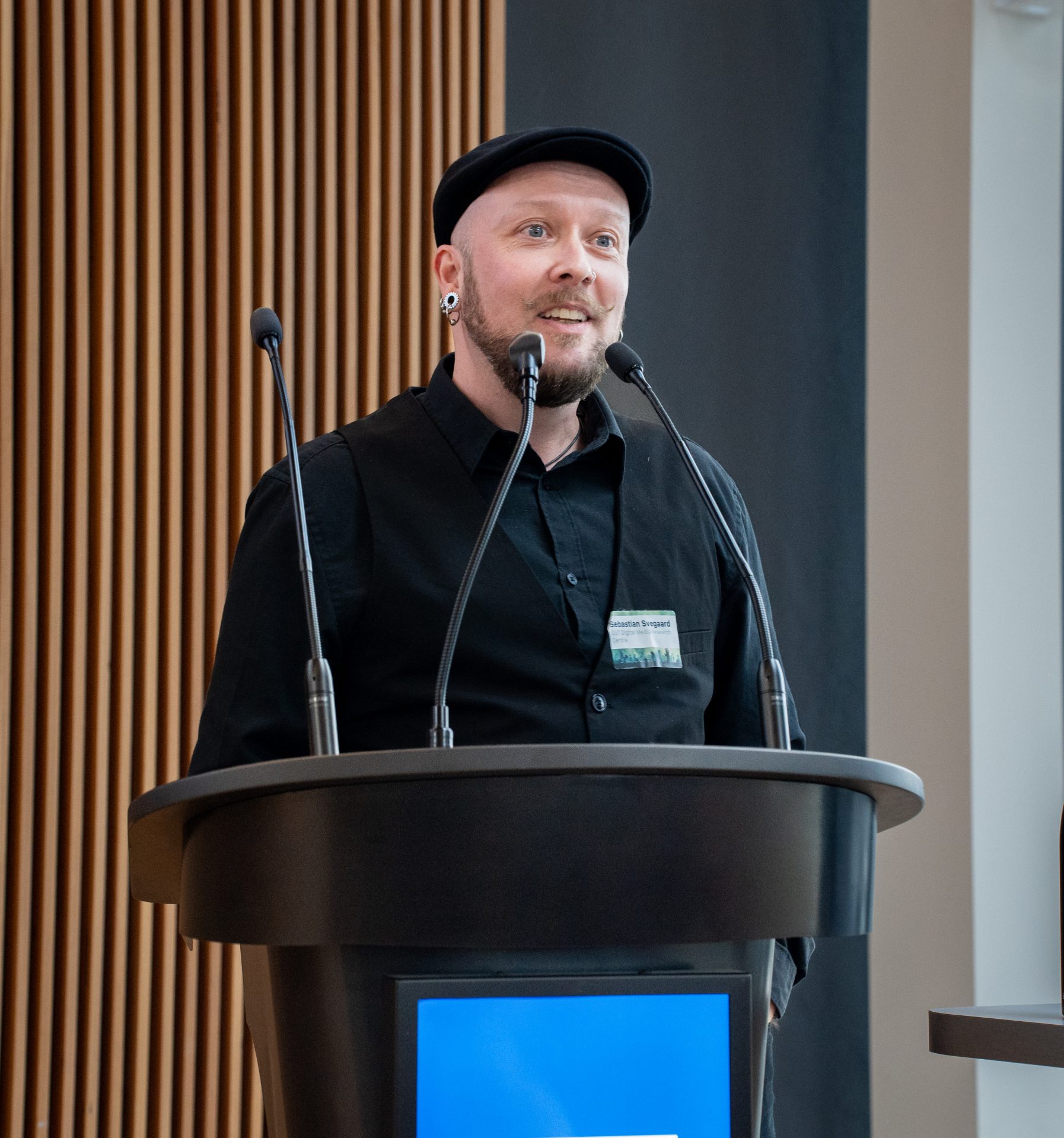 A man with a black cap and a black button-down shirt speaking into a microphone by a lectern.