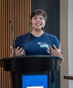 A woman with glasses and a blue shirt speaking into a microphone at a lectern.