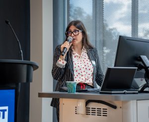 A woman with glasses and a patterned white button-down shirt speaking into a microphone by a lectern.