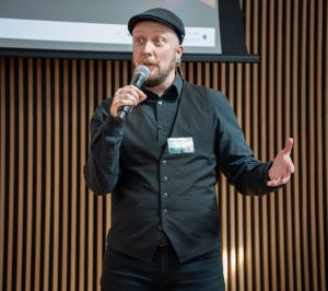 A man with a black cap and a black button-down shirt speaking into a microphone.