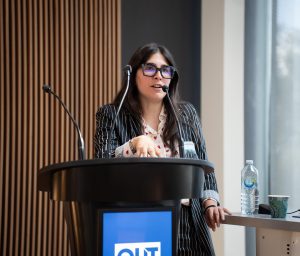 A woman with glasses and a patterned white button-down shirt speaking into a microphone by a lectern.