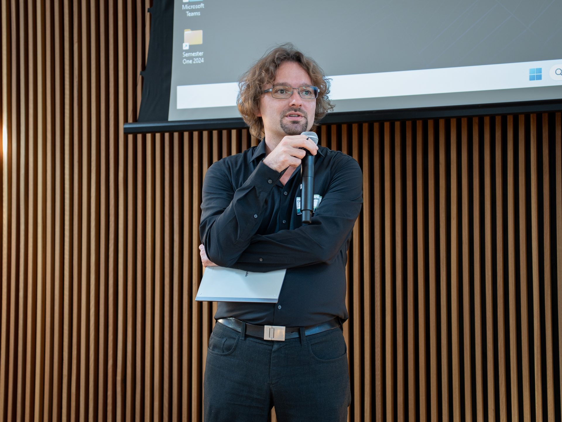 A man with glasses and a black button-down shirt with a microphone in one hand and a pamphlet in the other.