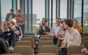 An audience with one member holding a microphone, asking a question. Two members seen conversing in the background.