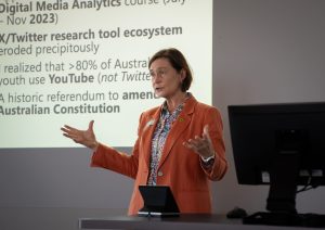 A woman wearing an orange coat gesturing while giving a presentation.