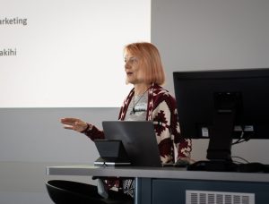 A woman wearing a patterned coat gesturing while giving a presentation.