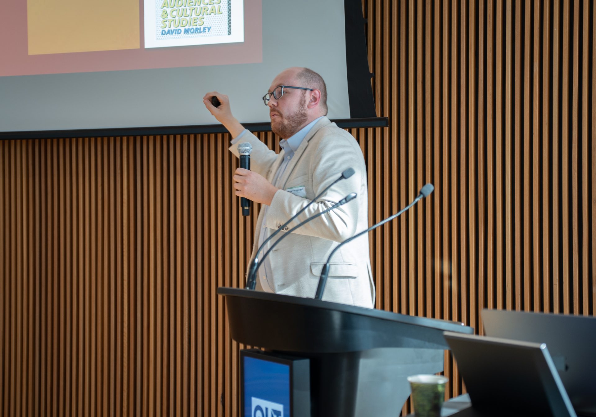 A man with glasses and a light beige coat gesturing towards a projection screen while speaking into a microphone and giving a presentation.