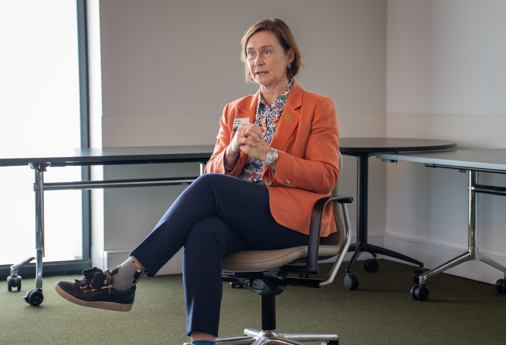 A woman in an orange coat sitting on a chair, holding her hands together while engaged in conversation with someone off-screen.