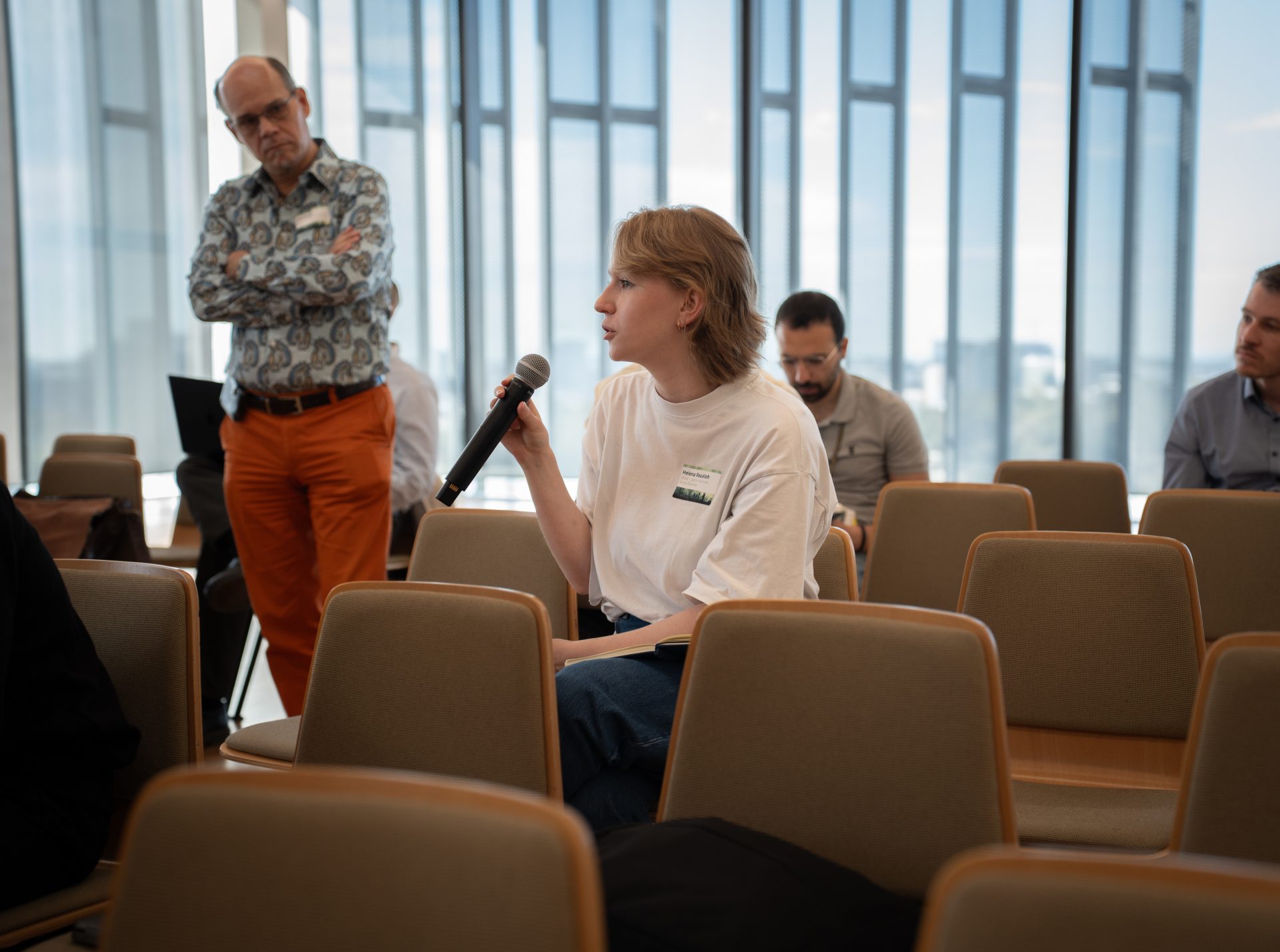 An audience with one member holding a microphone, another standing and looking towards the speaker, and others sitting in the background.