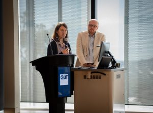 A man with glasses and a light beige coat, and a woman with straight hair and a black coat standing at a lectern, with the woman speaking into a microphone she is holding.