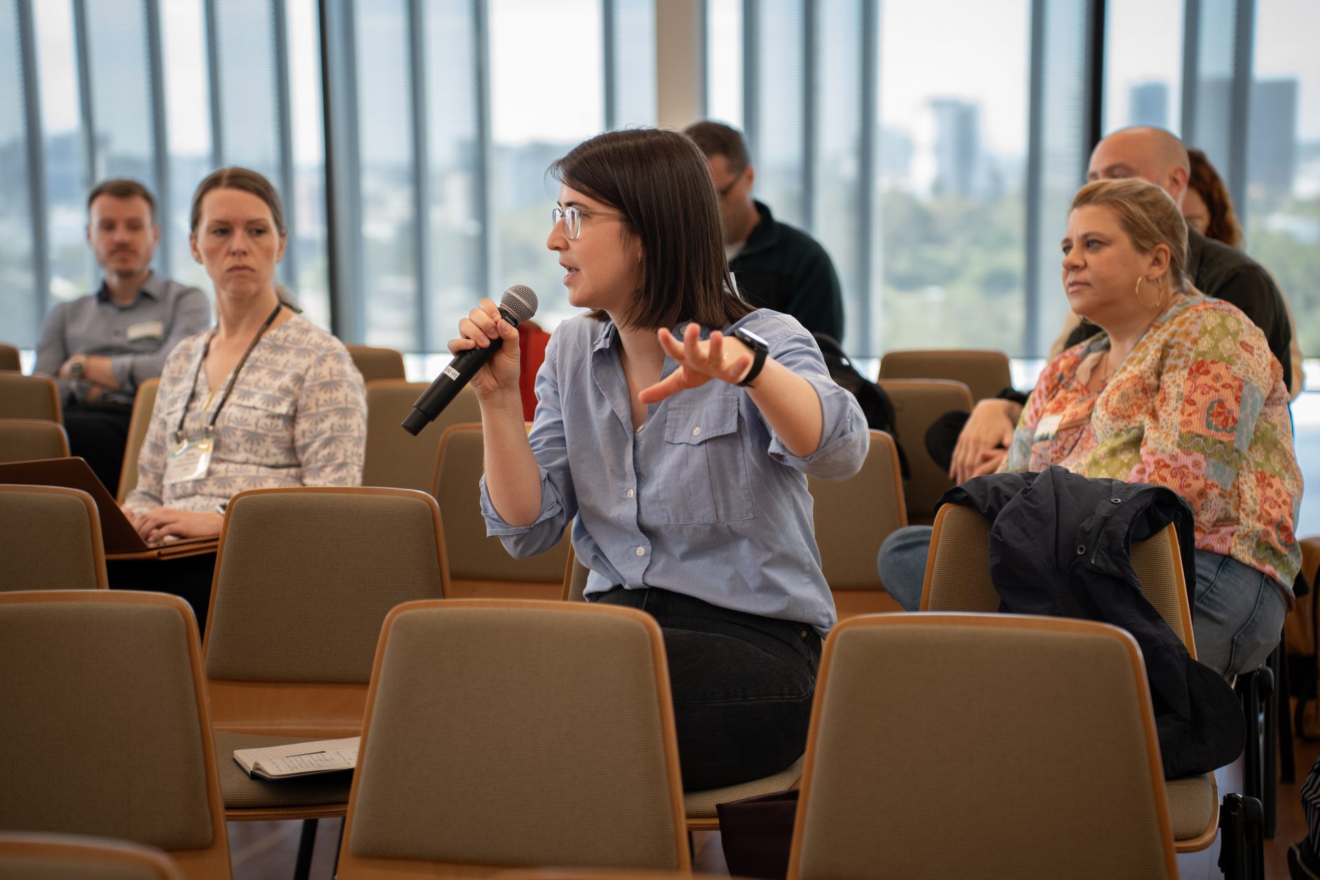 An audience with one member wearing a blue button-down shirt and straight hair, holding a microphone and speaking, while other audience members look at them.