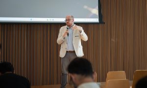 A man with glasses and a light beige coat holding a microphone and gesturing with his hands while speaking.