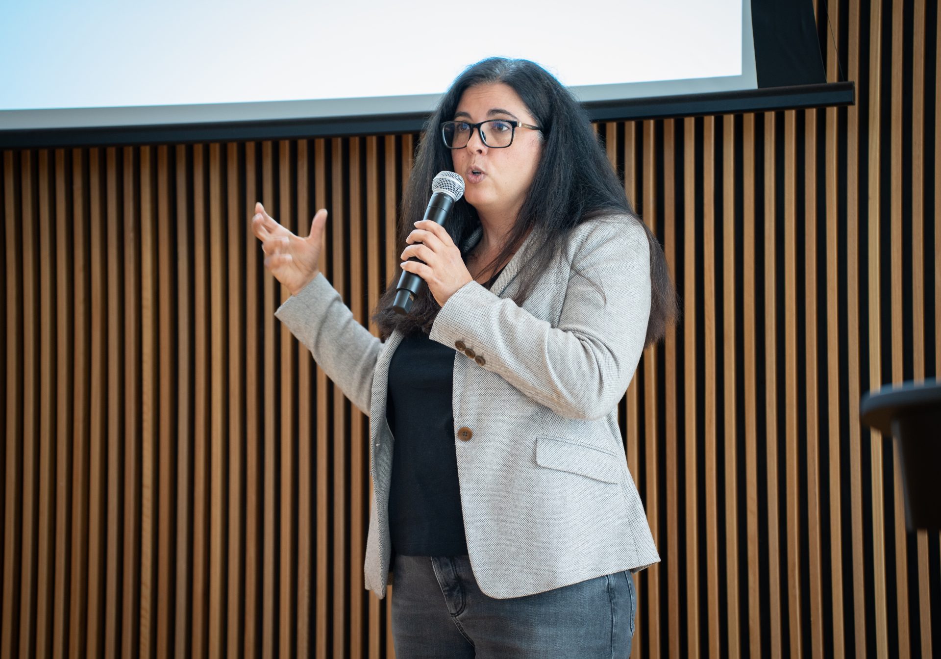 A woman with glasses and a light gray coat standing by a lectern holding a microphone and gesturing with her hands while speaking.