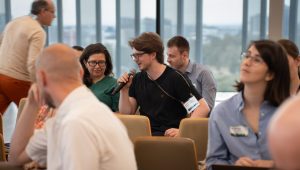 An audience with one member wearing a black shirt, holding a microphone and speaking, other audience members listening attentively.