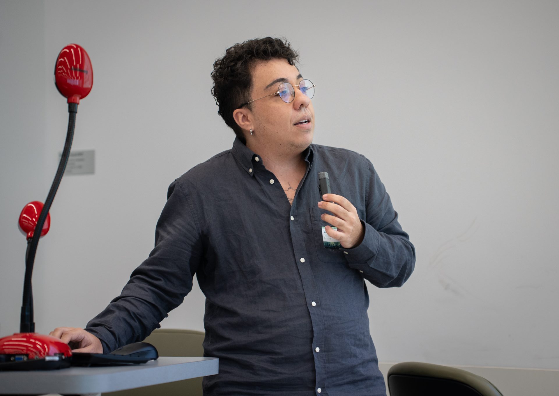 A man with curly hair and glasses, holding a microphone in one hand and resting his other hand on a table, addressing an audience.