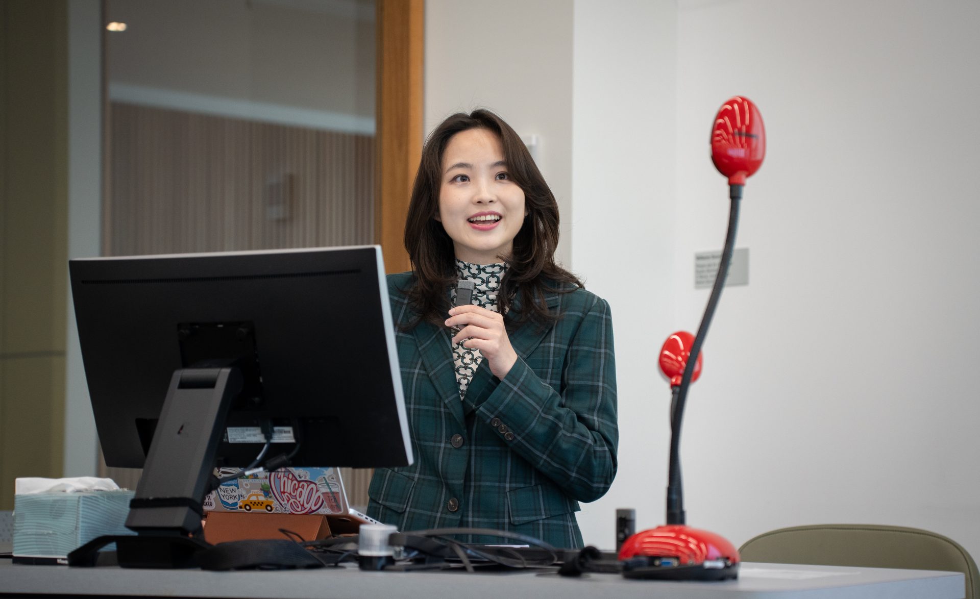 A woman with wavy hair and a green coat standing by a table with a monitor and a laptop, holding a microphone and speaking into it while addressing an audience.