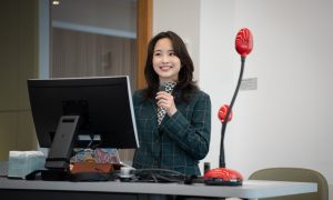 A woman with wavy hair and a green coat standing by a table with a monitor and a laptop, holding a microphone and speaking into it while addressing an audience.