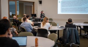 A presentation in progress at the front of a room, with audience members looking attentively at the screen.