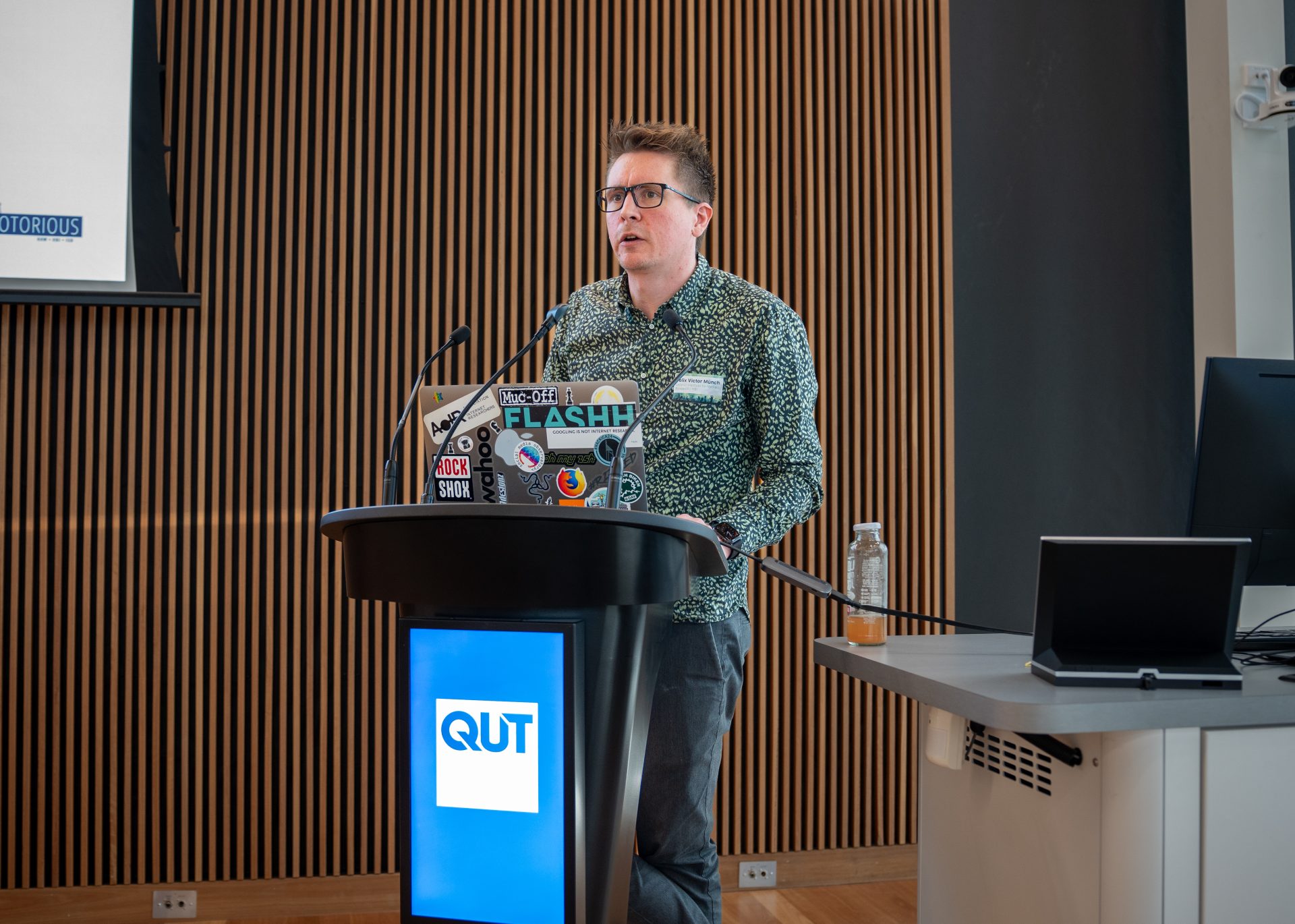 A man with glasses and a green patterned button-down standing by a lectern with a laptop, speaking into a microphone addressing an audience