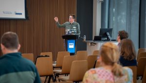 A presentation in progress at the front of a room, with audience members looking attentively at the screen. Presenter is gesturing with their hand towards the presentation.