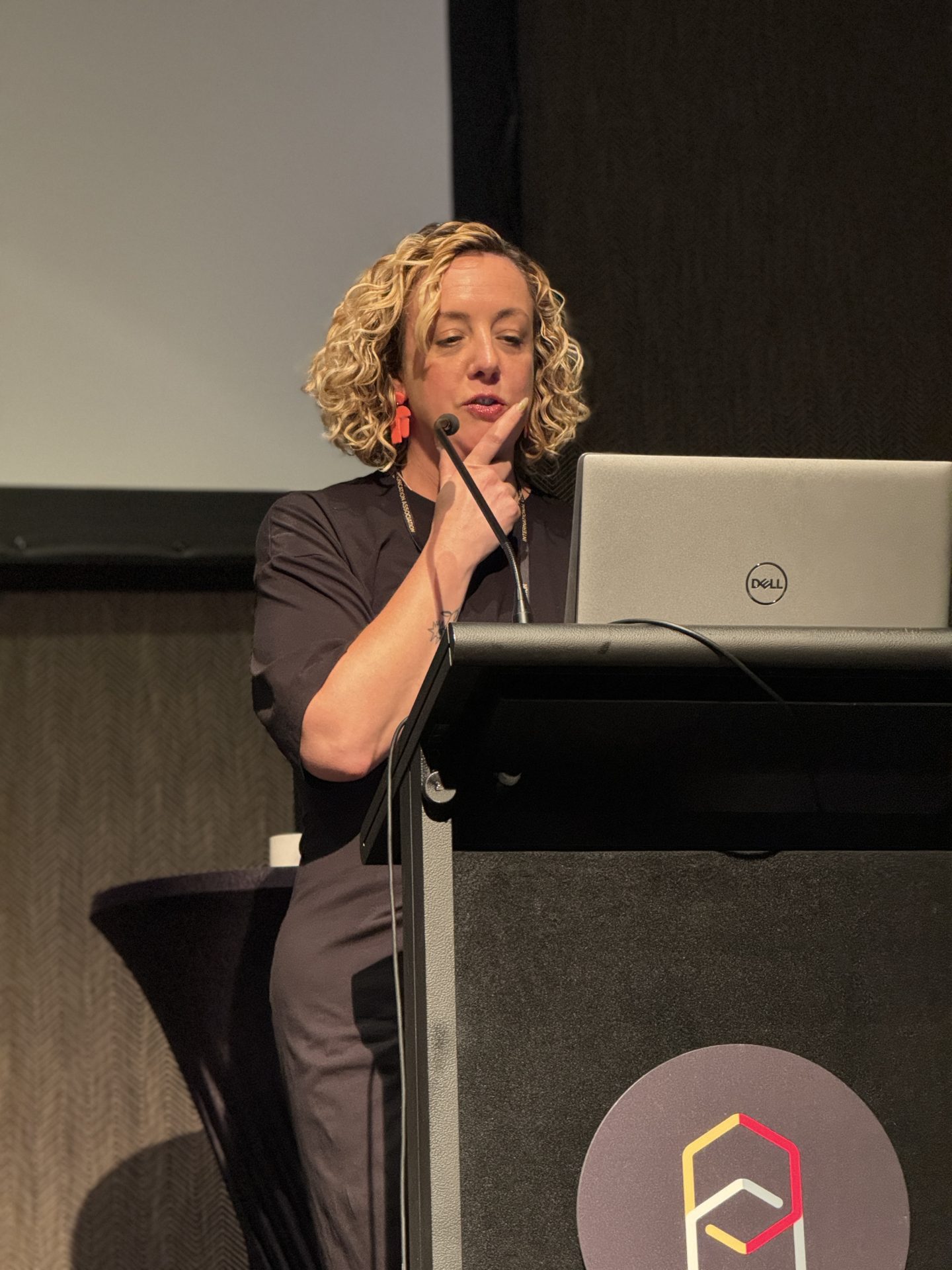 A woman with curly hair and a black dress stands by a lectern with a laptop speaking into a microphone, addressing an audience.