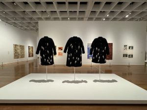 An art exhibition featuring three black coats on display, each adorned with small white figures. The coats are mounted on white stands in the center of a gallery with various paintings and artworks hanging on the walls in the background.