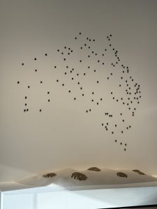 An art installation featuring a wall with numerous black dots arranged in a pattern resembling a map of Australia. Below the dots, there are three bronze-colored hand sculptures emerging from a white, smooth surface.