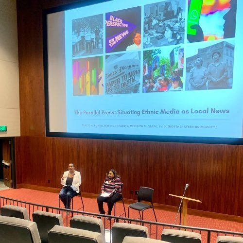 Meredith Clark and Tracie Powell sit in a large lecture hall beneath a large screen displaying the title of their talk, "The Parallel Press: Situating Ethnic Media as Local News"